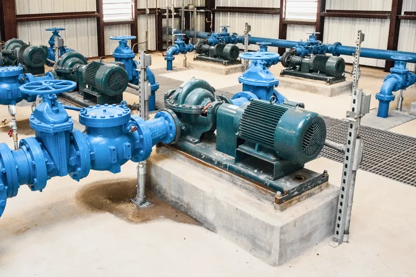 Casa de bombas com conjuntos de motor elétrico e bomba centrífuga sobre bases de concreto, ligados a uma tubulação azul com válvulas gaveta e de retenção