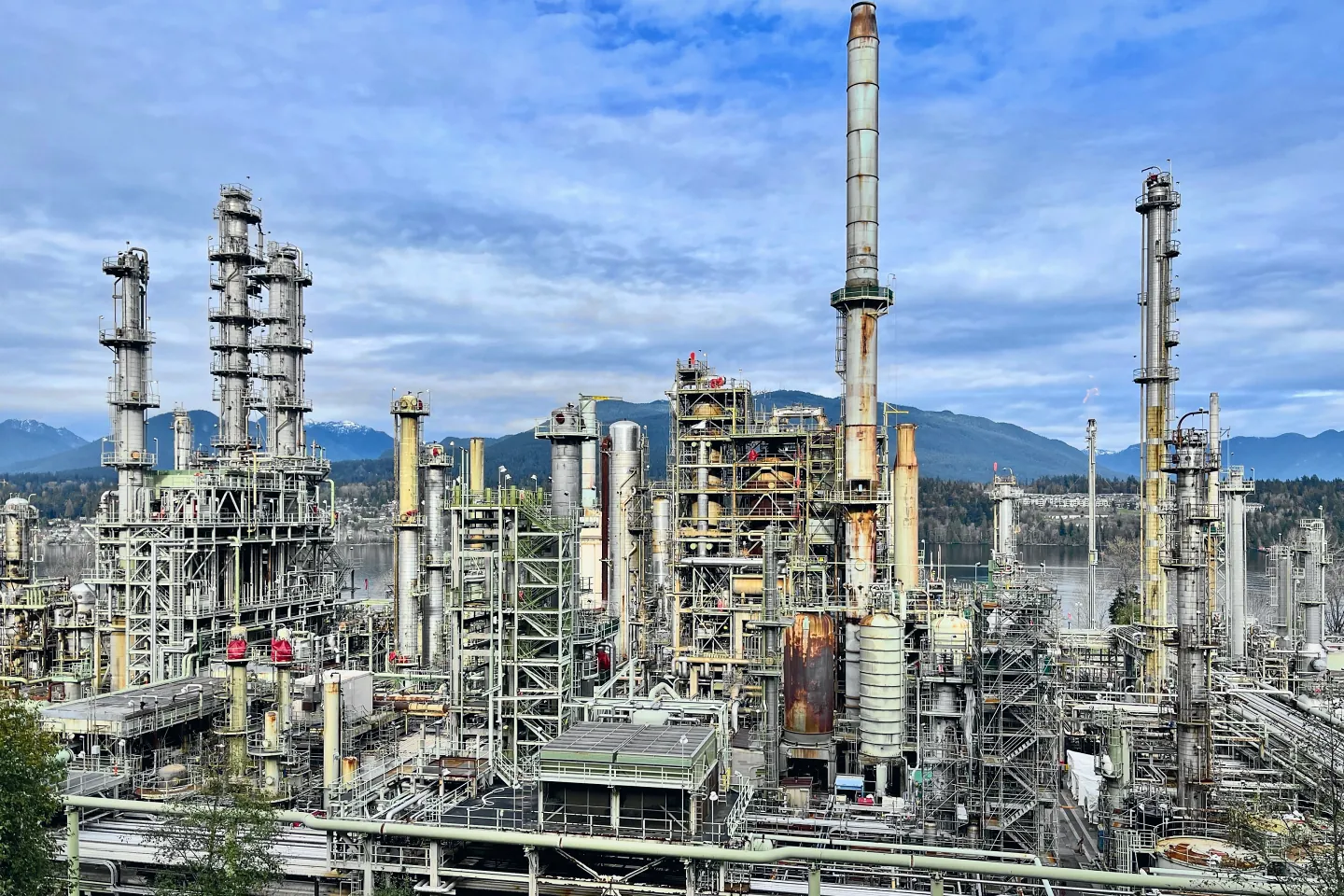 Refinaria em operação vista do alto de um talude: colunas de destilação com plataformas, vasos verticais, racks de tubulação e uma chaminé, com montanhas ao fundo