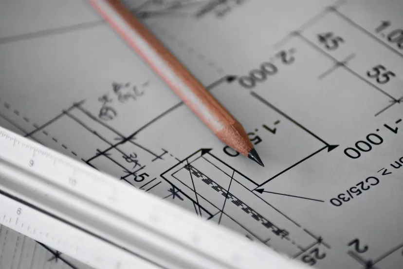 Lápis apontado sobre um desenho técnico com cotas e linhas de chamada, com um escalímetro de alumínio em primeiro plano